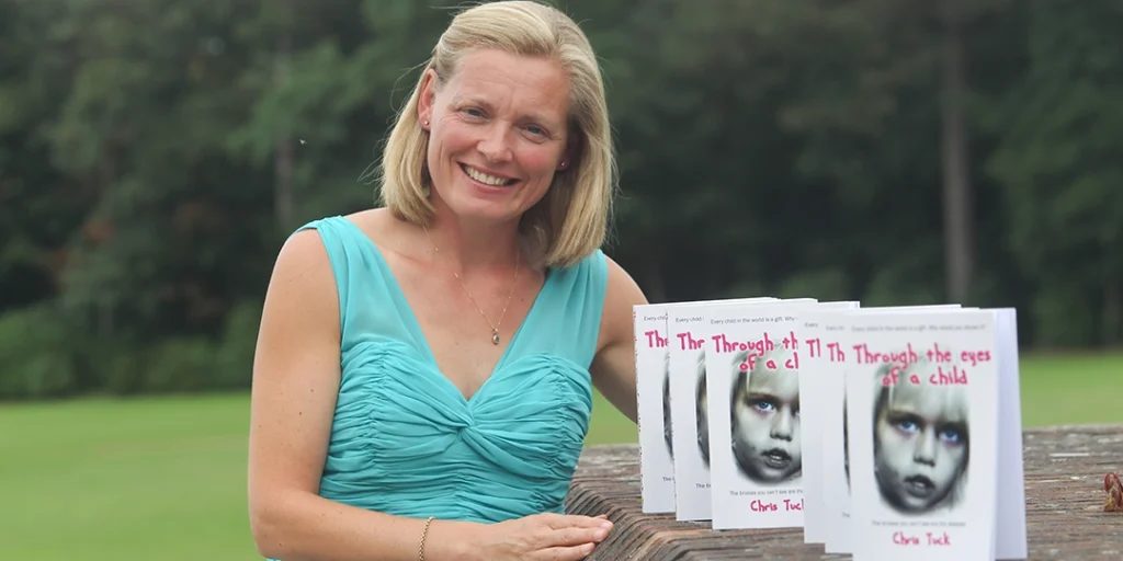 Author Chris Tuck smiling with her books 'Through the eyes of a child' standing on a wall